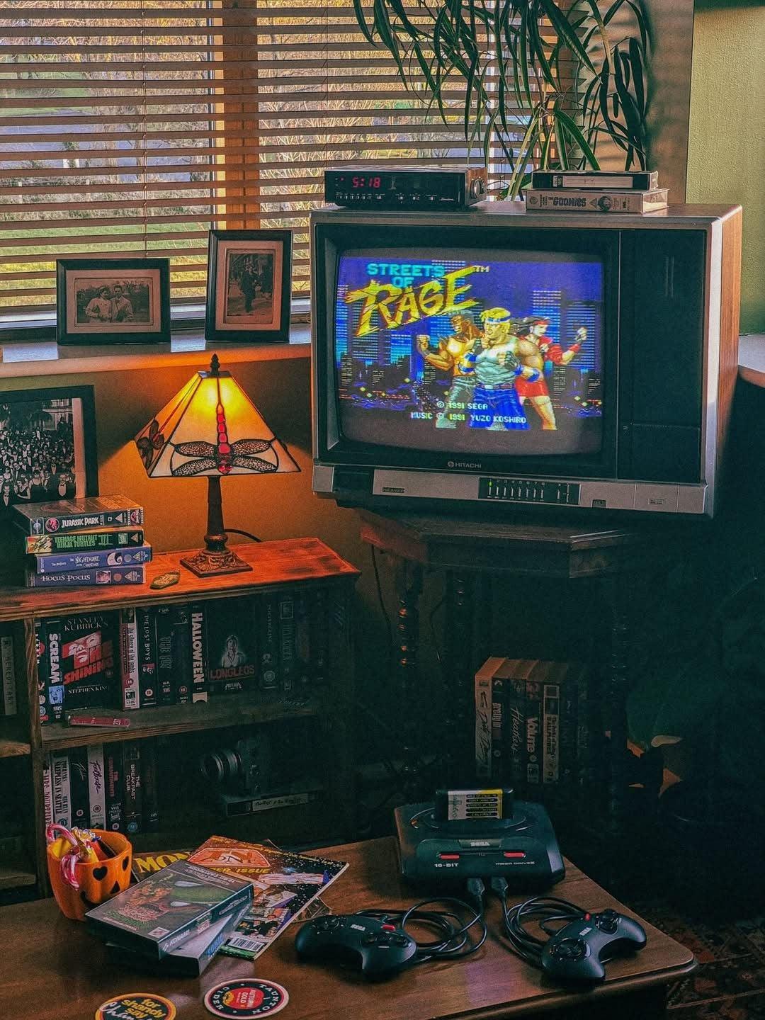 A retro 1980s CRT television displaying the game 'Street Rage' sits on a stand in a cozy, dimly lit room with bookshelves, a lamp, and a console table with controllers and game cases.