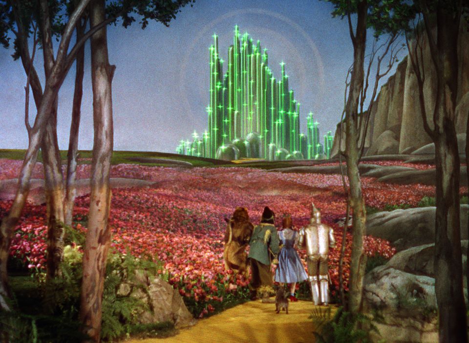 A scene from the 'Wizard of Oz' showing the four characters walking along a yellow path through a field of pink flowers toward a glowing green castle under a blue sky, framed by trees.